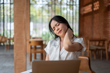 Fototapeta premium Tired asian woman holding or touching stiffed neck in front of laptop on table while sitting in cafe