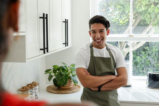 African American man smiling and chatting in home kitchen wearing olive-green apron, near plant