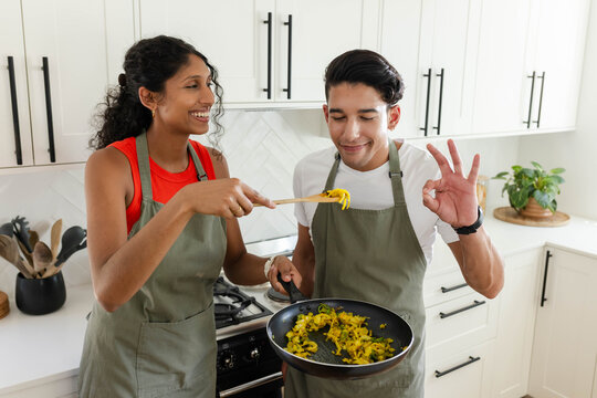 African American couple cooking on gas range in home kitchen in olive aprons, sampling wooden spoon