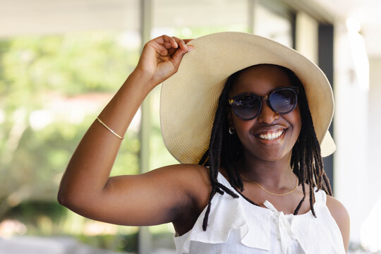 Woman smiling, holding straw hat standing on porch in white ruffled top and sunglasses, copy space