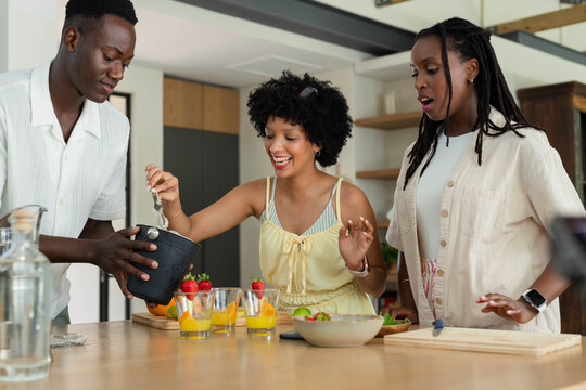 African American friends scooping fruit into glasses at wooden kitchen island with spoon