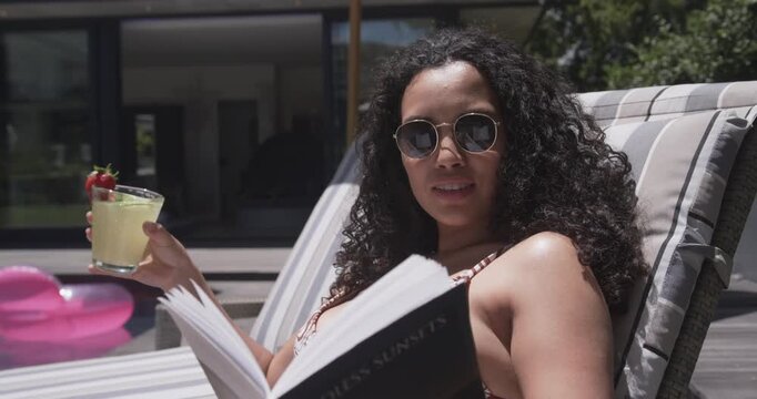Adult woman reclining by pool zebra bikini, holding book and drink, smiling as camera moving closer