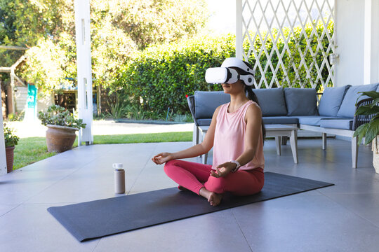 Woman is sitting cross-legged on yoga mat on tiled patio wearing white VR headset, bottle visible