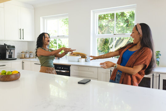 Two women are standing opposite white kitchen island, pointing, laughing near phone and citrus bowl