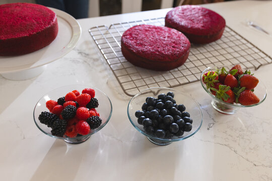 Three glass pedestal bowls are sitting across white marble countertop with berries and cake layers
