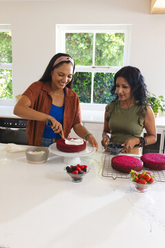Diverse women frosting cake using offset spatula on turntable and adding berries at home kitchen