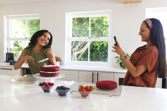 Diverse female friends frosting red velvet cake with offset spatula and filming on phone on counter