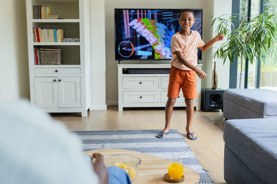 African American parent and boy standing watching colorful TV in living room, boy wearing peach tee