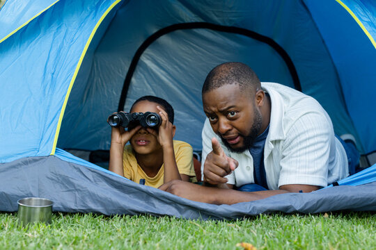 African American dad, child lying at blue tent on grass with metal cup, using binoculars, pointing