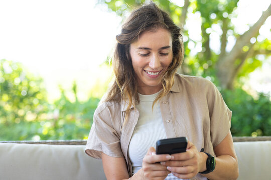 Woman sitting on patio sofa holding black smartphone and smiling, wearing beige overshirt