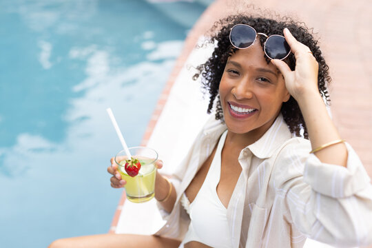 Woman sitting poolside wearing white bikini and open shirt with drink, lifting shades, copy space