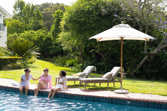 Diverse female friends wearing swimwear dipping feet in pool, chatting under white umbrella