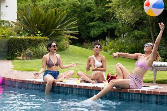 Diverse female friends sitting in swimwear on brick coping at home pool, tossing beach ball