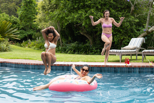 Female friends in bikinis with shades laughing, jumping into backyard pool near pink donut float