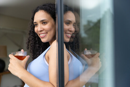 African American woman holding clear glass cup with amber drink near glass door reflection