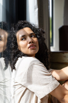 Adult African American woman sitting by window with reflection wearing plain tee near sill, curtain