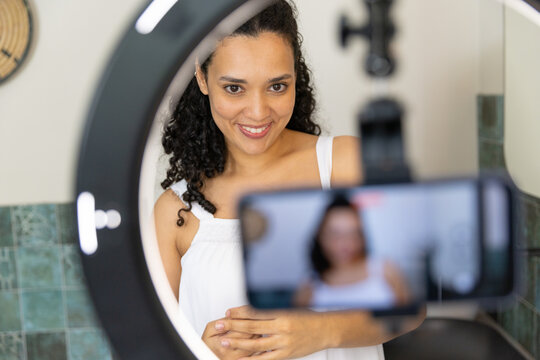 African American woman filming at vanity under ring light wearing white top with phone tripod