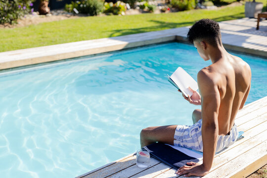 African American man sitting on pool deck reading book shirtless in trunks towel jar, copy space