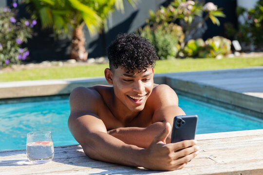 African American adult man shirtless leaning on wood pool deck, holding phone and glass with citrus