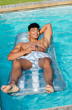 African American man floating on pool float in pool in blue-striped trunks holding glass