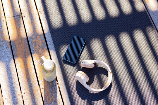 Light headphones are resting on mat on weathered deck with smartphone and bottle, casting stripes
