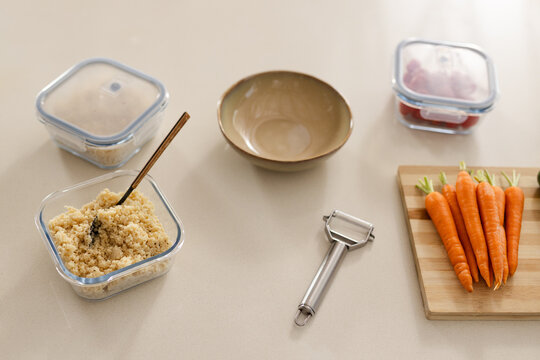 Square glass container is holding cooked grain with wooden spoon on countertop, carrots nearby