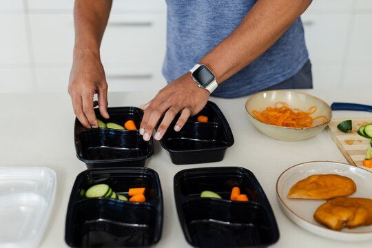 Four black plastic meal-prep containers being filled with cucumber rounds, carrots on white counter