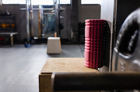 Pink foam roller is leaning vertically in gym on plyo box near barbell shaft and rack