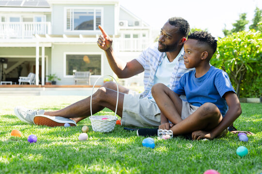 African American father and teenage son sitting on lawn, pointing toward basket with eggs