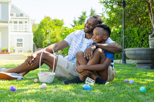 Mature father and youth son sitting on front lawn smiling while holding basket with pastel eggs