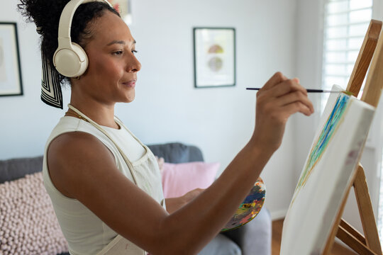 African American woman painting at wooden easel in living room, wearing apron and holding palette