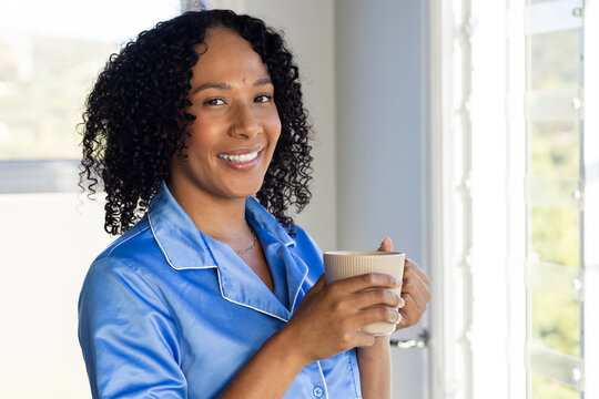 Woman standing by shuttered window holding ceramic mug smiling in blue pajama top and gold necklace
