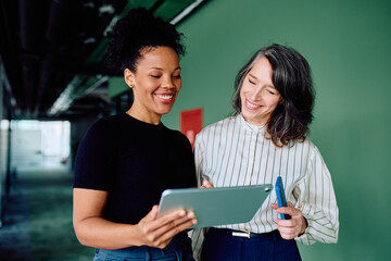 Two diverse businesswomen discussing work and laughing while using a digital tablet for collaboration and technology in a modern office