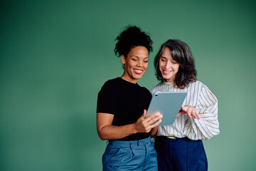 Two smiling businesswomen from different backgrounds are sharing a digital tablet, discussing and...
