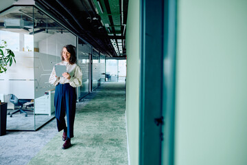Businesswoman walking through a contemporary office corridor, smiling and engrossed in using a digital tablet