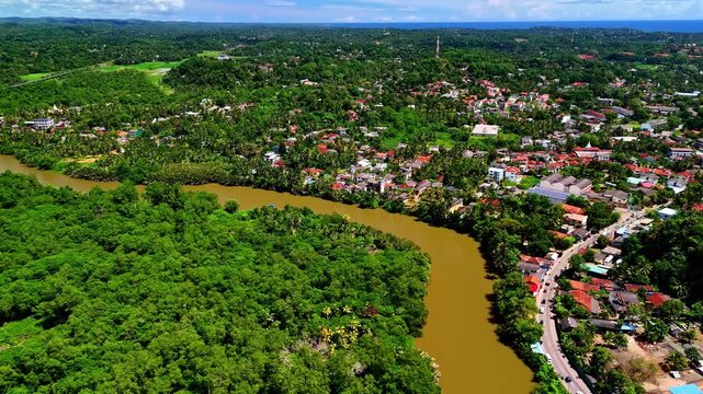 An aerial drone view of Matara's Nilwala River shows muddy water winding between dense greenery and a bustling town with houses, roads, and shops along its banks in Sri Lanka.