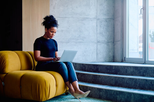 Concentrating businesswoman using a laptop while sitting on a yellow designer sofa in a modern office space, remote work concept