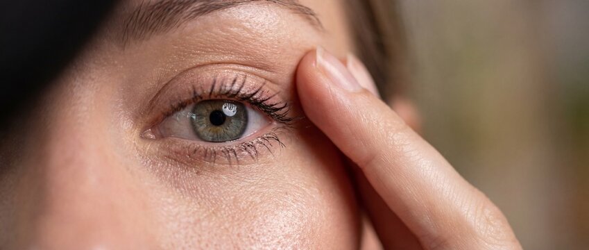 Close-up of a Caucasian woman's green eye and hand gently touching the upper eyelid, symbolizing beauty, skincare, ophthalmology, and self-care routine.