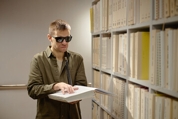 Young adult Caucasian man wearing sunglasses reading braille book with hands standing in library...