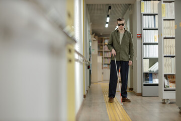Fototapeta premium Young adult Caucasian man wearing sunglasses navigating library hallway using cane, following tactile guidance path, demonstrating independent mobility for visually impaired