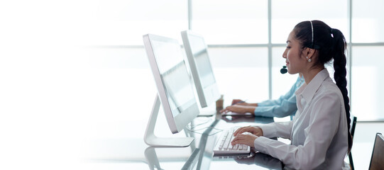 Fototapeta premium Customer service representative wearing headset working at computer in modern office. Call center support agent providing online assistance, communication and professional service.