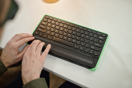 Caucasian middle aged man using braille display keyboard with both hands on desk in library setting, demonstrating assistive technology for blind individuals accessing information