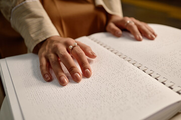Young adult woman reading braille book with both hands in library, focusing on tactile text,...