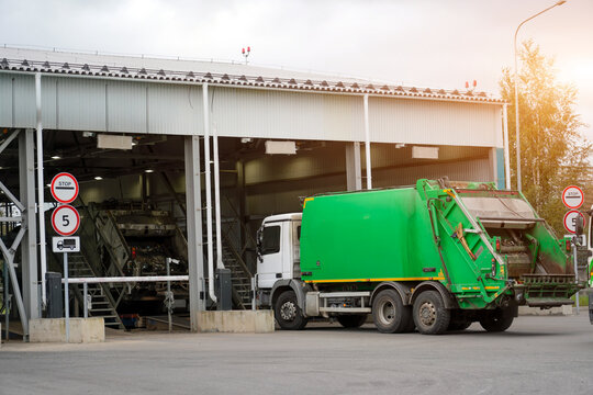 Bright green refuse truck waits at the entrance gate of a large waste processing facility. Inside the industrial building, another vehicle unloads collected garbage. Sunlight illuminates the scene.