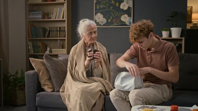 Teenager unzipping small bag and taking blister of pills from it while sitting on couch next to sick grandmother wrapped in blanket drinking hot tea and looking at grandson