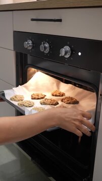A woman is taking cookies out of an oven. The cookies are on a baking sheet and are ready to be eaten
