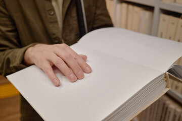 Obraz premium Middle aged Caucasian man reading braille book by touching raised dots with left hand in library, shelves with books visible in background, focusing on tactile literacy for blind individuals