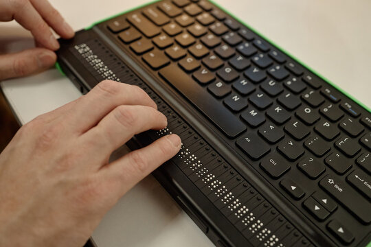 Caucasian adult man using fingers to read braille display on electronic keyboard in library setting, demonstrating assistive technology for blind individuals accessing information