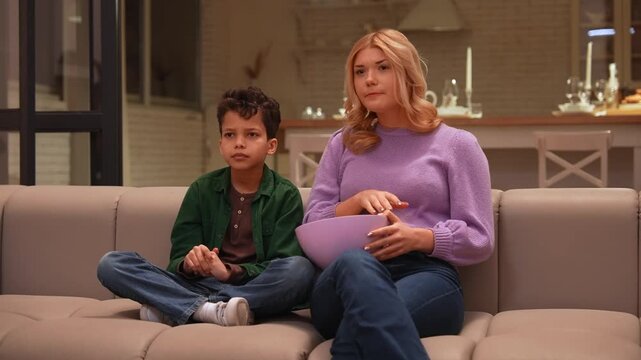 Charming teenage babysitter protecting adorable African American school age boy from adult content on tv, covering child eyes with hand and continuing to watch with interest indoors.