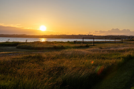 Golden sunset near Ebeltoft, Denmark, with warm evening light over coastal meadows and tall grass, the sun reflecting on the water in the background, and football goal frames silhouetted against the h